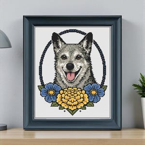 May include: Framed cross-stitch art featuring a smiling dog with gray and white fur, a pink tongue, and a black collar. The dog is surrounded by blue and yellow floral accents, set against a white background. The frame is dark blue.