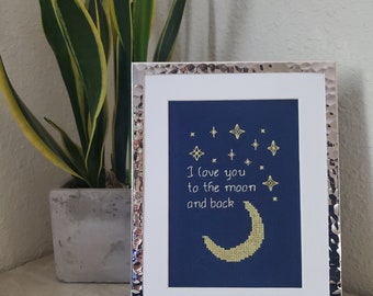 Pode incluir: Um bordado de ponto cruz emoldurado com um fundo azul marinho e bordado dourado. O bordado representa uma lua crescente e estrelas com o texto "Eu te amo até a lua e de volta".