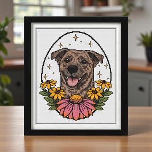 May include: Framed cross-stitch art featuring a smiling brown dog with its tongue out, surrounded by yellow and pink flowers. The dog is set against a white background with an oval border and star accents. The frame is black.