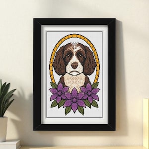 Op de afbeelding: Een ingelijste kruissteekafbeelding van een hond met bruin en wit haar, geplaatst in een gouden ovaal. Paarse bloemen met groene bladeren sieren de onderkant. Het kunstwerk zit in een zwarte lijst en staat op een houten oppervlak.