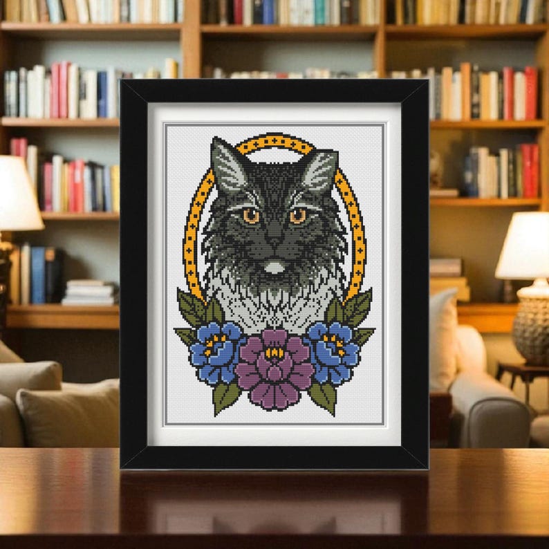 Op de afbeelding: Een ingelijst kruissteekkunstwerk met een zwarte kat met oranje ogen, omringd door een gele, gestippelde ovaal en bloemmotieven. Het kunstwerk is ingelijst in een zwarte lijst.