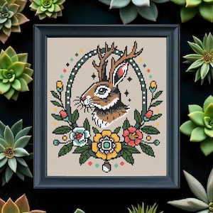 Framed jackalope cross stitch pattern with antlers and colorful tattoo-style flowers, surrounded by green succulents for a modern rustic display.