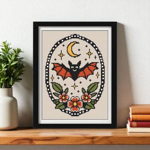 Peut inclure: Une œuvre d'art au point de croix encadrée représentant une chauve-souris noire aux ailes orange, un croissant de lune jaune et des étoiles. La chauve-souris est entourée de fleurs rouges et de feuilles vertes, le tout dans un cadre ovale.