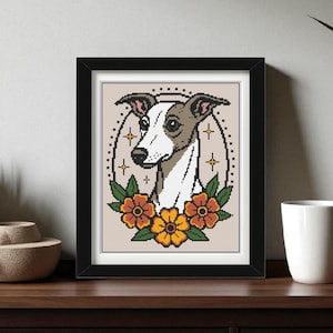 May include: Framed cross-stitch art depicting a greyhound dog. The dog has a white face and grey ears, encircled by orange and yellow flowers. The artwork is in a black frame, displayed on a wooden surface.
