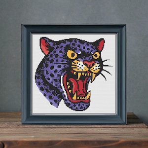 Puede incluir: Obra de arte de punto de cruz enmarcada que representa una cabeza de pantera rugiente. La pantera es morada con manchas negras, orejas rojas y boca roja abierta. Los ojos son amarillos. El marco es azul oscuro y está sobre madera.