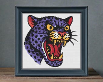 May include: Framed cross-stitch art featuring a snarling panther head. The panther is purple with black spots, red ears, and a red open mouth revealing white teeth. The eyes are yellow. The frame is dark blue, and the artwork is displayed on a wooden surface.