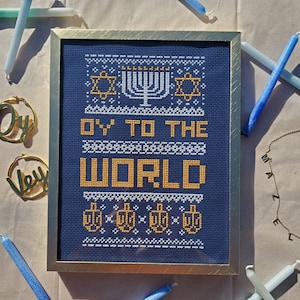 May include: A framed cross-stitch design on blue fabric with white and gold thread. The design features a menorah, stars of David, and the text "Oy to the World".