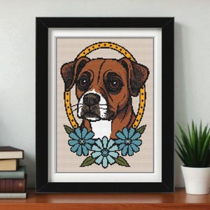 May include: Framed cross-stitch art featuring a Boxer dog portrait. The dog has brown fur, dark eyes, and a white muzzle. A yellow halo surrounds the dog's head, and blue flowers are at the bottom. The artwork is in a black frame.