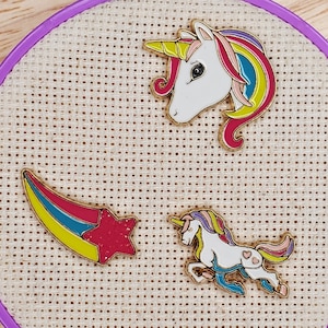May include: Three colorful enamel pins featuring a unicorn, a unicorn running, and a shooting star. The pins are on a beige fabric background.