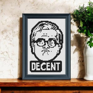 Może przedstawiać: Oprawiony portret haftem krzyżykowym w czerni i bieli, przedstawiający osobę w okularach i słowo "DECENT" poniżej. Rama jest granatowa, a grafika jest wyświetlana na drewnianej powierzchni.