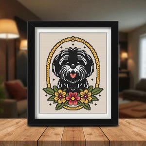 May include: Framed cross-stitch art featuring a black and white dog within a gold oval frame, surrounded by flowers. The dog has a pink tongue. The artwork is displayed in a black frame, set on a wooden surface.