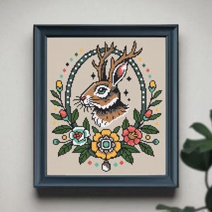 Framed jackalope cross stitch pattern with antlers and bold tattoo-style flowers, displayed on a neutral wall with a leafy plant in the corner.