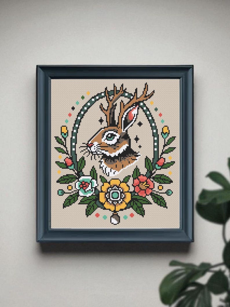Framed jackalope cross stitch pattern with antlers and bold tattoo-style flowers, displayed on a neutral wall with a leafy plant in the corner.