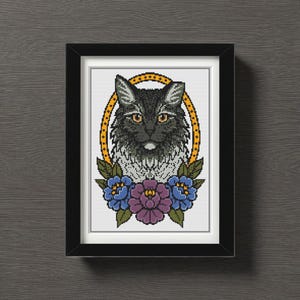 Op de afbeelding: Een ingelijste kruissteekkunst met een gedetailleerd portret van een zwarte kat met gele ogen, geplaatst in een gouden ovaal. Het ontwerp bevat blauwe en paarse bloemaccenten en is ingelijst in een zwart rechthoekig frame.