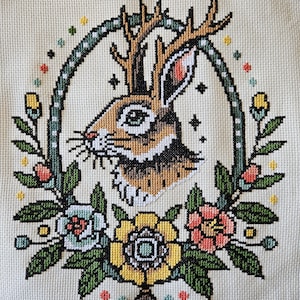 Jackalope cross stitch pattern featuring a rabbit with antlers surrounded by colorful tattoo-style flowers and leaves on beige fabric.