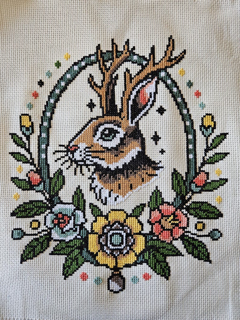 Jackalope cross stitch pattern featuring a rabbit with antlers surrounded by colorful tattoo-style flowers and leaves on beige fabric.