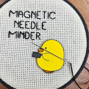 May include: A yellow chick needle minder with a black magnetic holder. The text "MAGNETIC NEEDLE MINER" is embroidered on a white canvas.