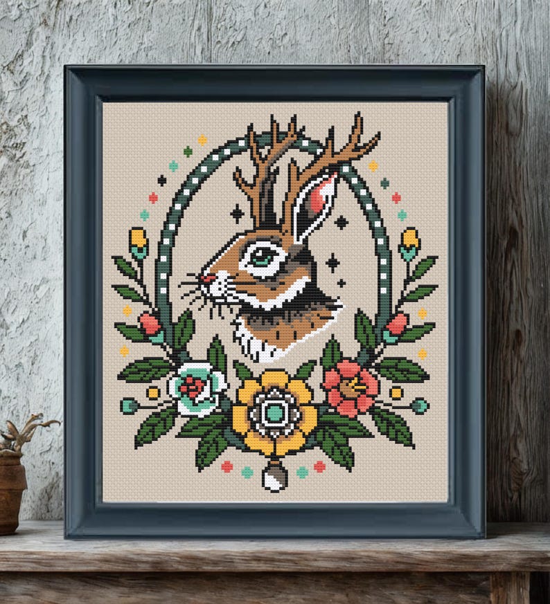 Framed jackalope cross stitch pattern with antlers and colorful traditional tattoo-style flowers, displayed on a rustic wooden surface against a textured wall.
