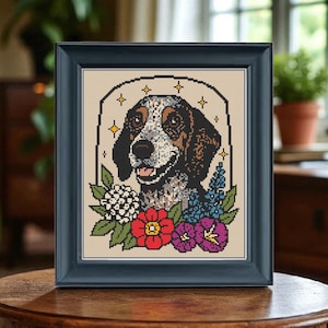 以下が含まれることがあります： 茶色と白の模様の犬とカラフルな花が描かれた、額入りのクロスステッチアート。 作品はベージュの背景に設定され、濃い青色のフレームに収められています。 犬は幸せそうな表情をしています。