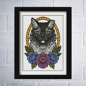 Op de afbeelding: Ingelijste kruissteekkunst met een zwarte kat met gele ogen, omringd door een gouden ovaal en bloemaccenten. De bloemen zijn blauw en paars. Het kunstwerk wordt gepresenteerd in een zwart frame tegen een witte bakstenen muur.