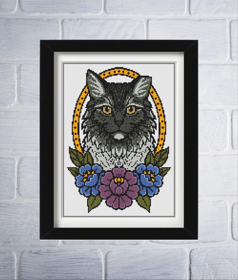 Op de afbeelding: Ingelijste kruissteekkunst met een zwarte kat met gele ogen, omringd door een gouden ovaal en bloemaccenten. De bloemen zijn blauw en paars. Het kunstwerk wordt gepresenteerd in een zwart frame tegen een witte bakstenen muur.
