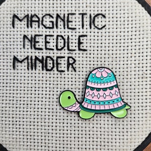 May include: A green and pink turtle-shaped magnetic needle minder with the text "MAGNETIC NEEDLE MINDER" embroidered on a beige fabric background.