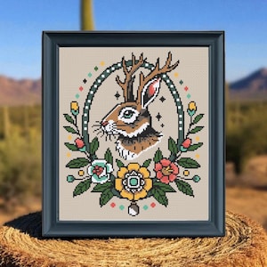 Framed jackalope cross stitch with antlers and colorful tattoo-style flowers displayed outdoors on a hay bale with a desert cactus background.