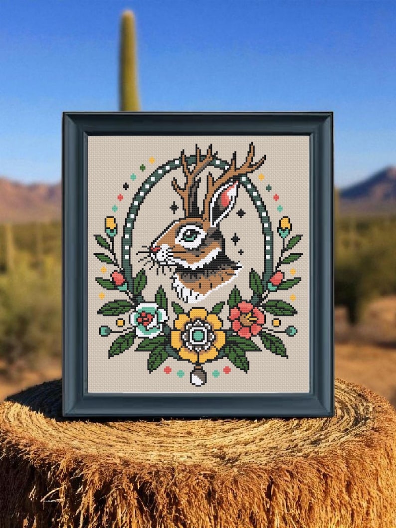 Framed jackalope cross stitch with antlers and colorful tattoo-style flowers displayed outdoors on a hay bale with a desert cactus background.