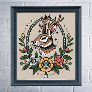 A cross-stitch pattern of a brown and white jackalope inside of a tall oval frame with white dots. There are multicolored flowers along the base of the frame and dots of color along the outside.