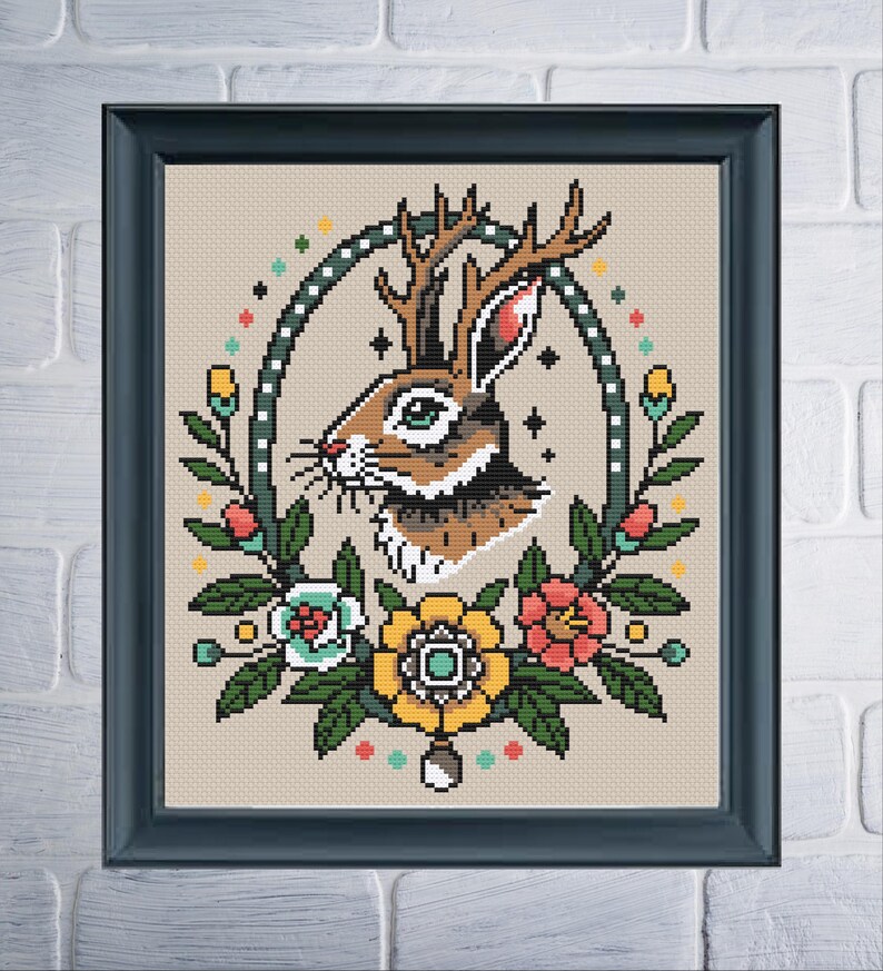 A cross-stitch pattern of a brown and white jackalope inside of a tall oval frame with white dots. There are multicolored flowers along the base of the frame and dots of color along the outside.