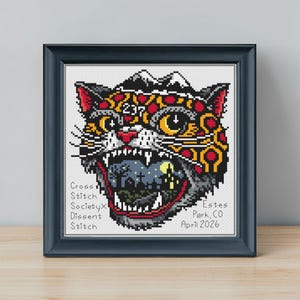 May include: A framed cross stitch artwork featuring a stylized jaguar head with a mountain peak on top, the number 237, and a graveyard scene inside its open mouth. Text reads Cross Stitch SocietyX Dissent Stitch, Estes Park, CO, April 2026.