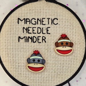 May include: A close-up of a beige fabric circle with the text "Magnetic Needle Minder" embroidered in black. Two small, magnetic monkey needle minders are attached to the fabric. One monkey is red, white, and black, and the other is blue, white, and black.