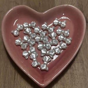 Könnte beinhalten: Eine Ansammlung kleiner, perlmuttfarbener, weißer Schleifenverzierungen, die in einer herzförmigen, rosa Keramikschale angeordnet sind. Die Schleifen haben eine glänzende Oberfläche und sind über die Schale verstreut, was eine zarte und charmante Ästhetik erzeugt.