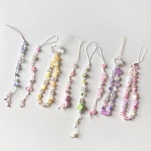 Puede incluir: Colección de siete colgantes de teléfono con cuentas en varios colores pastel. Cada colgante presenta un diseño diferente, incluyendo estrellas, conchas y lazos. Los colgantes están ensartados en cordones blancos.