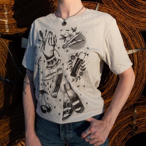 Puede incluir: Una camiseta blanca con un estampado gráfico en blanco y negro de varios artículos, incluyendo una mano, un cortaúñas, una cereza, un cuchillo, una botella de esmalte de uñas y un par de sandalias.