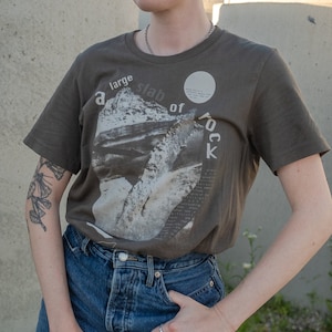 Puede incluir: Una persona lleva una camiseta marrón con un estampado gráfico de una gran losa de roca. El texto "a large slab of rock" está impreso en la camiseta. La persona lleva pantalones vaqueros azules y tiene un tatuaje en el brazo.
