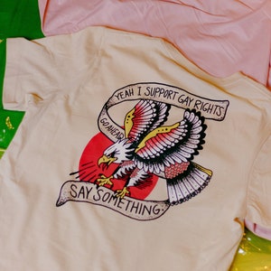 Puede incluir: Una camiseta blanca con un gráfico de un águila roja, blanca y azul. El águila sostiene una pancarta que dice "Yeah I support gay rights go ahead say something".