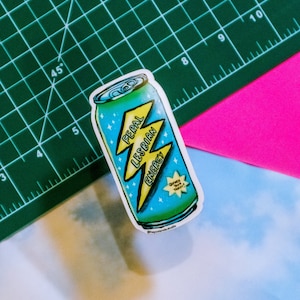 Puede incluir: Una pegatina de una lata azul y amarilla con un rayo y el texto "Feral Lesbian Energy". La lata tiene una estrella blanca y el texto "Quench your thirst!" en ella.