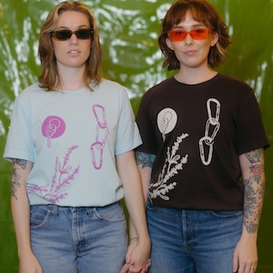 Puede incluir: Dos personas con camisetas con un gráfico de una flor, una cadena y un cono de helado. La persona de la izquierda lleva una camiseta azul claro y la persona de la derecha lleva una camiseta negra. Ambas personas llevan gafas de sol.