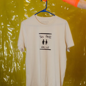 Puede incluir: Una camiseta de color crema con texto negro que dice "two they's" y una ilustración de dos personas tomadas de la mano. El texto debajo de la ilustración dice "PMS.ng".