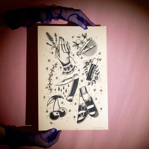 Puede incluir: Una impresión artística de tatuajes en blanco y negro con varios diseños de tatuajes tradicionales. La impresión incluye manos rezando, una navaja de bolsillo, cerezas, un tubo de protector labial y otros motivos clásicos de tatuajes. La obra de arte está sostenida por una persona con guantes azules.