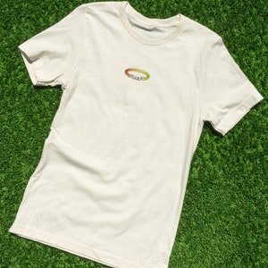 Puede incluir: Una camiseta blanca con un pequeño diseño de pulsera de cuentas de colores en la parte delantera. La pulsera está hecha de pequeñas cuentas redondas en tonos de rojo, naranja, amarillo y verde. Las cuentas están dispuestas en un círculo y deletrean la palabra "JUICY".