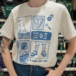 Puede incluir: Una camiseta blanca con un estampado gráfico azul de una cortadora de césped, un filete, una tuerca y un tornillo, un control remoto, un par de sandalias y dos tapas de botellas. El texto "be your own dad" está impreso debajo del gráfico.