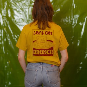 Puede incluir: Una persona lleva una camiseta amarilla con un gráfico rojo que dice "Let's Get Brunch" y una pila de panqueques. La persona lleva pantalones vaqueros azules.