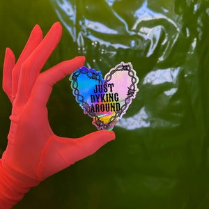 Puede incluir: Una pegatina holográfica en forma de corazón con alambre de púas alrededor de los bordes. La pegatina dice "Just Dyking Around" en texto negro.
