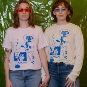 Puede incluir: Dos personas con camisetas rosa claro y crema con un diseño floral azul y el texto "season it's pansy season". Llevan vaqueros azules y gafas de sol. El fondo es una superficie verde texturizada.