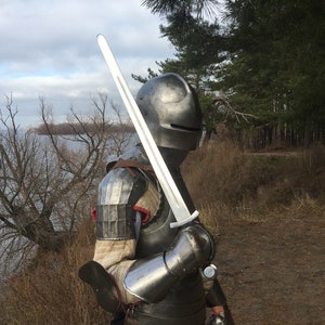 May include: A knight in full armor stands with a sword in hand, facing away from the camera. The knight is wearing a helmet with a visor, and the sword is held at a diagonal angle. The knight is standing on a grassy hill, with a body of water in the background.