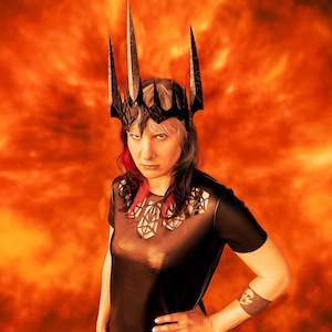 Peut inclure: Une couronne noire avec trois pointes hautes et pointues. La couronne est portée par une personne portant un haut noir à manches courtes avec un motif géométrique découpé au niveau de l'encolure. Le fond est orange flamboyant.