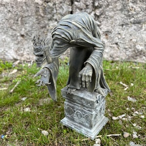 May include: A bronze-colored statue of a cloaked figure with a crown, leaning forward on a pedestal. The figure has a detailed face and outstretched hand. The pedestal has ornate carvings. The statue is set on a grassy surface.