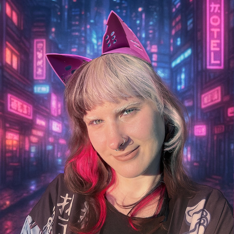May include: A person wearing a pink cat ear headband with cut-out details. The person is smiling, with a nose ring and pink and white hair. The background features a neon-lit cityscape with Japanese text.